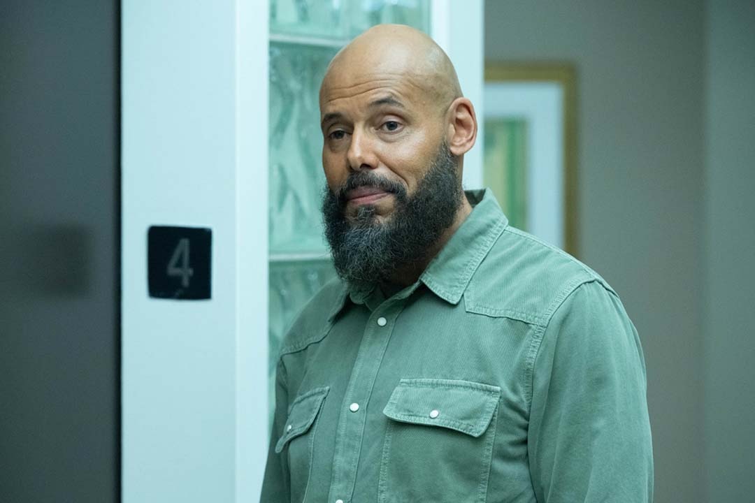 August Ripley (Maurice Dean Wint) stands in a green shirt near an elevator in SurrealEstate 205.