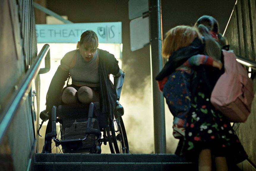 Fiona Dourif as Nica Pierce (Fiona Dourif) appears in a wheelchair at the top of the stairs as a little girl holding Chucky looks up at her in Chucky 303.