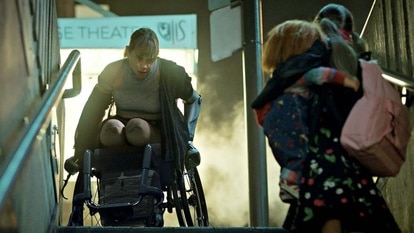 Fiona Dourif as Nica Pierce (Fiona Dourif) appears in a wheelchair at the top of the stairs as a little girl holding Chucky looks up at her in Chucky 303.